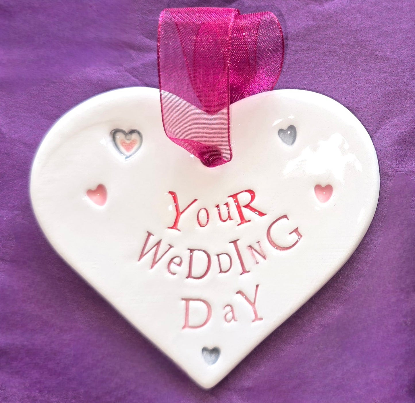 White ceramic heart with the words "Your Wedding Day" and some small heart illustrations dotted around. There is a burgundy ribbon at the top of the heart for hanging.