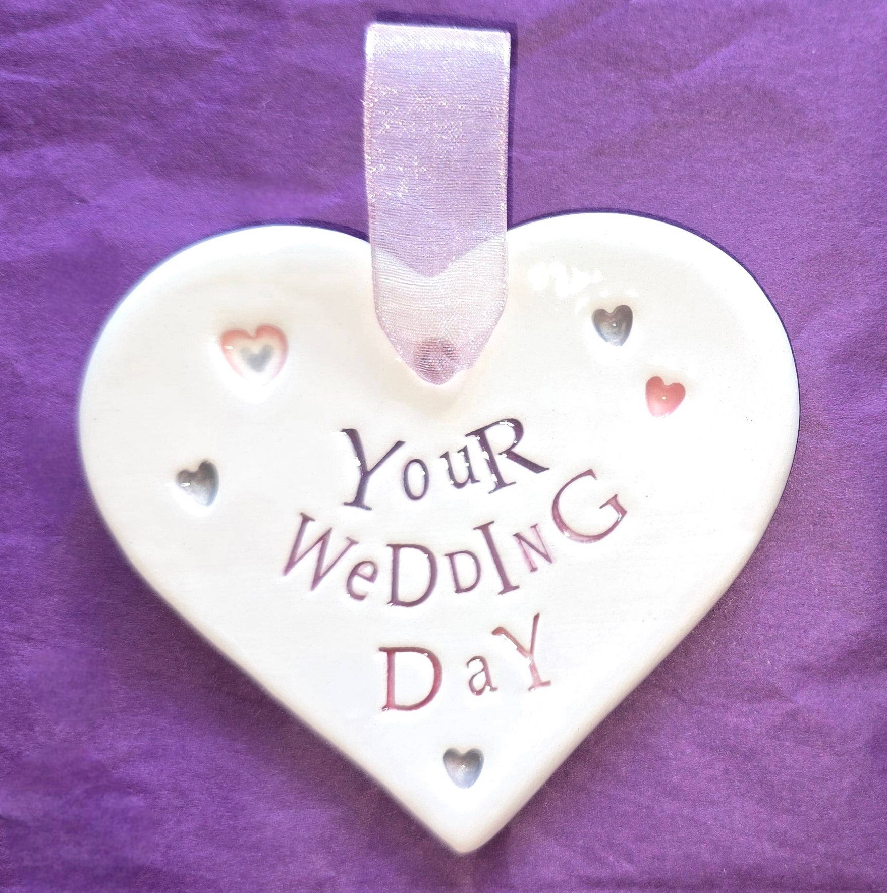 White ceramic heart with the words "Your Wedding Day" and some small heart illustrations dotted around. There is a pale pink ribbon at the top of the heart for hanging.