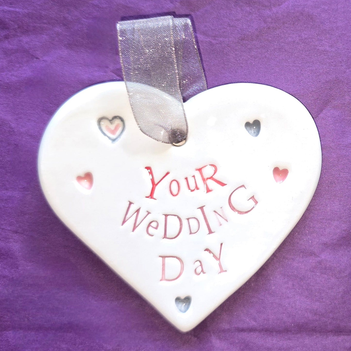 White ceramic heart with the words "Your Wedding Day" and some small heart illustrations dotted around. There is a silver ribbon at the top of the heart for hanging.