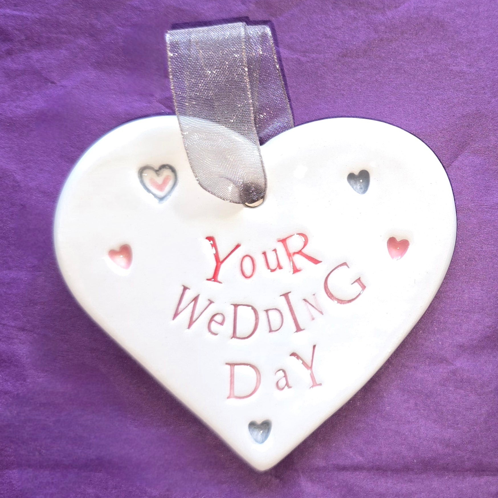 White ceramic heart with the words "Your Wedding Day" and some small heart illustrations dotted around. There is a silver ribbon at the top of the heart for hanging.